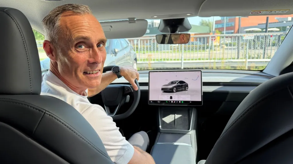 Marc Diks lachend op de bestuurdersstoel van een Tesla, kijkend over zijn schouder vanaf het stuur, met het centrale FSD-touchscreen in beeld.